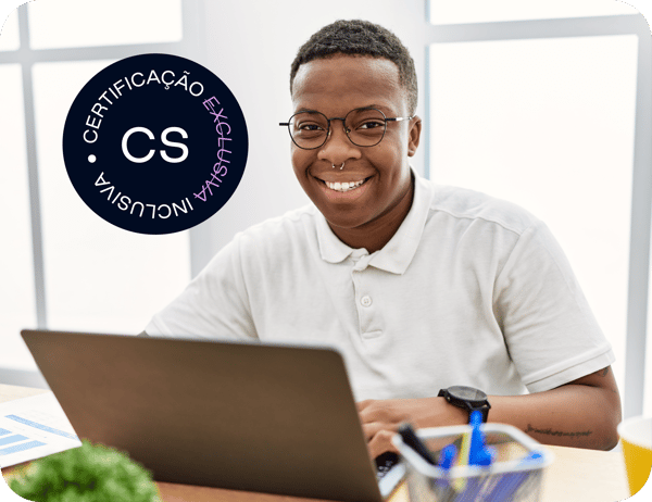 Homem negro sorrindo, usa óculos, camisa branca e um relógio no braço esquerdo. Está sentando de frente para uma mesa com um notebook e canetas.