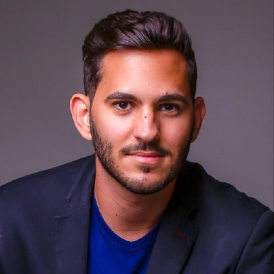 Homem de pele parda com cabelo castanho escuro e curto, penteado para trás. Veste uma camiseta azul e um blazer preto e está sorrindo para a câmera.