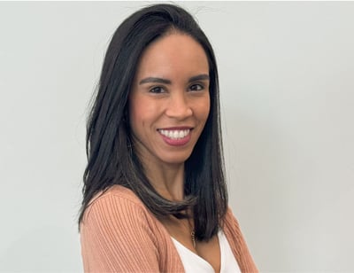  Mulher de pele parda, cabelos lisos e pretos, usando blusa branca e cardigan salmão. Ela está sorrindo com os braços cruzados.