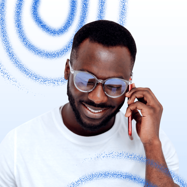 Homem de pele preta, barba, óculos de grau e camiseta branca. Ele está falando ao telefone com um leve sorriso no rosto.